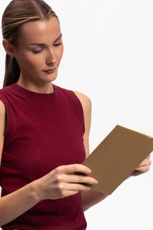 Young woman in sleeveless red top reads a book against a white background. Focused expression and natural lighting highlight concentration and casual style in a simple studio setting.
