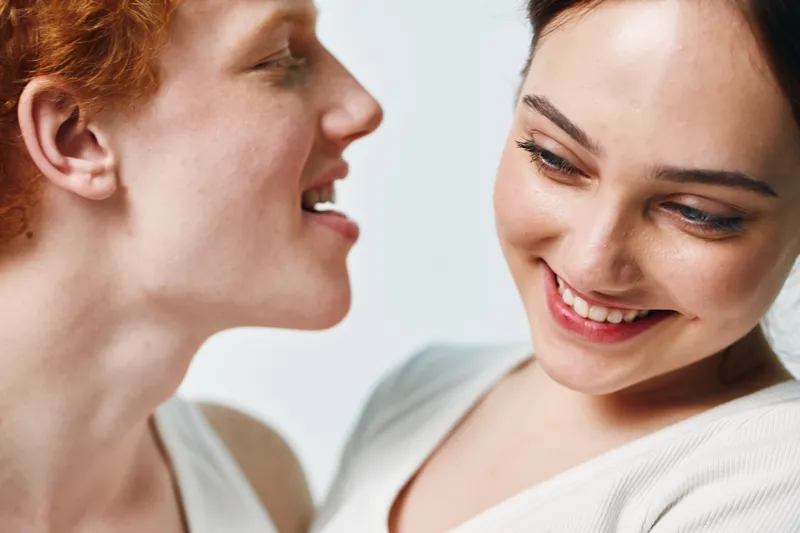 Two women smiling and interacting closely with joyful expressions in a bright environment, displaying friendship and positive emotions in a candid moment.