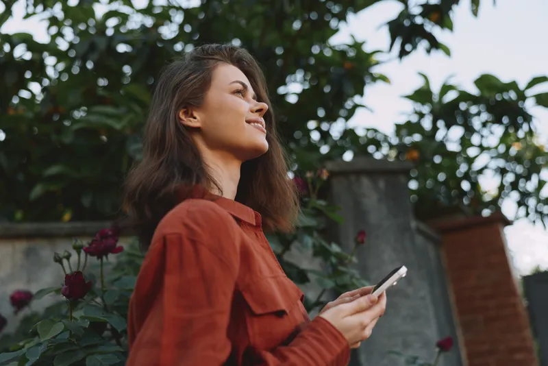Smiling woman in a red shirt outdoors, looking up with a smartphone in hand. Natural light highlights a casual lifestyle and warm garden ambiance