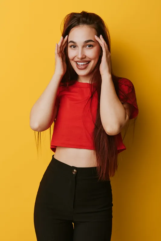 Young woman with long hair wearing red crop top and black pants smiling and touching face standing against yellow background in casual style portrait with happy expression.
