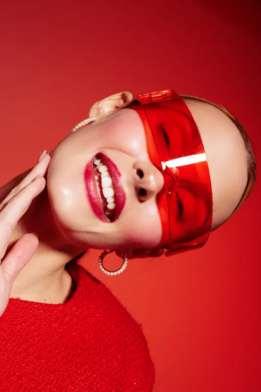 Smiling woman with red transparent glasses and red sweater poses on red background. Close-up portrait with earrings and bright makeup. Concept of fashion, style, and confidence.