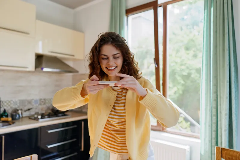 Joyful woman at home in a bright kitchen, smiling while taking a photo with her phone, casual outfit, everyday life scene, intimate domestic mood, cheerful and relaxed.
