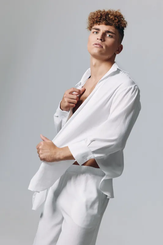 Young man with curly hair wearing loose white shirt and pants posing confidently against plain background in studio for fashion or lifestyle portrait.