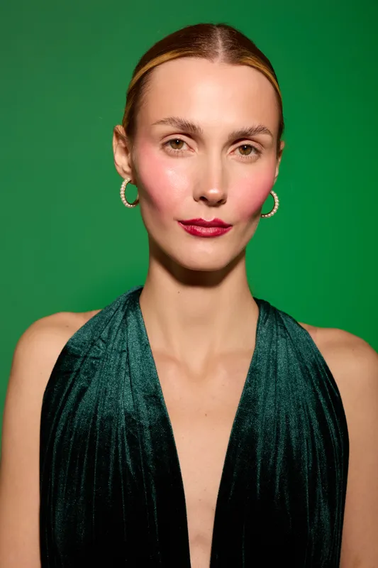 Portrait of elegant woman with slicked hair wearing green velvet dress and gold hoop earrings against green background. Concept of fashion, beauty, and confident female model.
