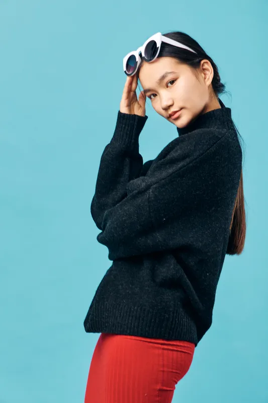 Young woman wearing black sweater and red skirt poses with white sunglasses on head against blue background. Fashion model standing with side glance and confident expression.