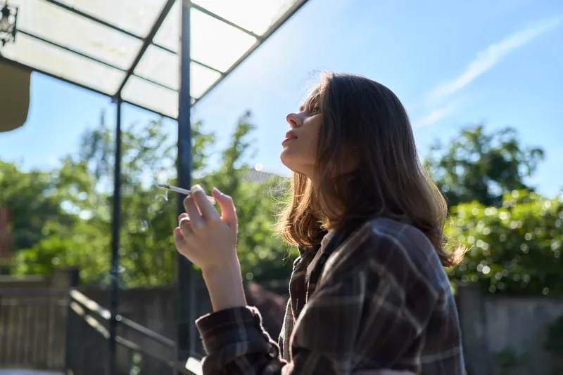 Woman lifestyle portrait outdoors in sunlight, wearing plaid shirt, looking up with a relaxed thoughtful expression in a garden setting