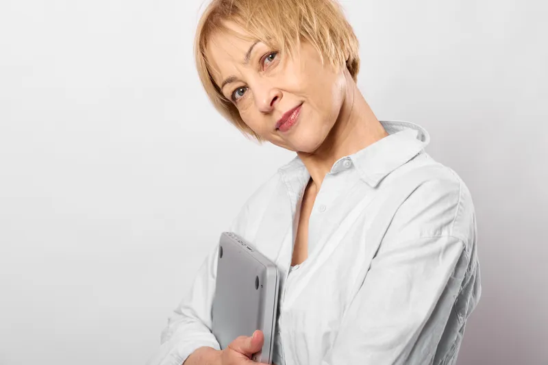 Senior woman with short blond hair holding a smartphone, gentle smile in studio portrait, white blouse on light background, technology concept, calm mood