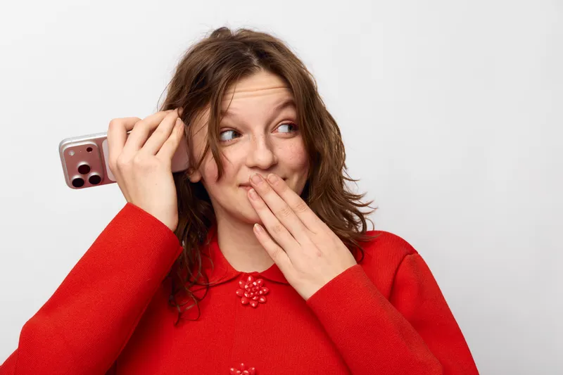 Young woman in red sweater holds smartphone near ear and covers mouth with hand, looking surprised and playful. Concept of communication, secret, and casual lifestyle.