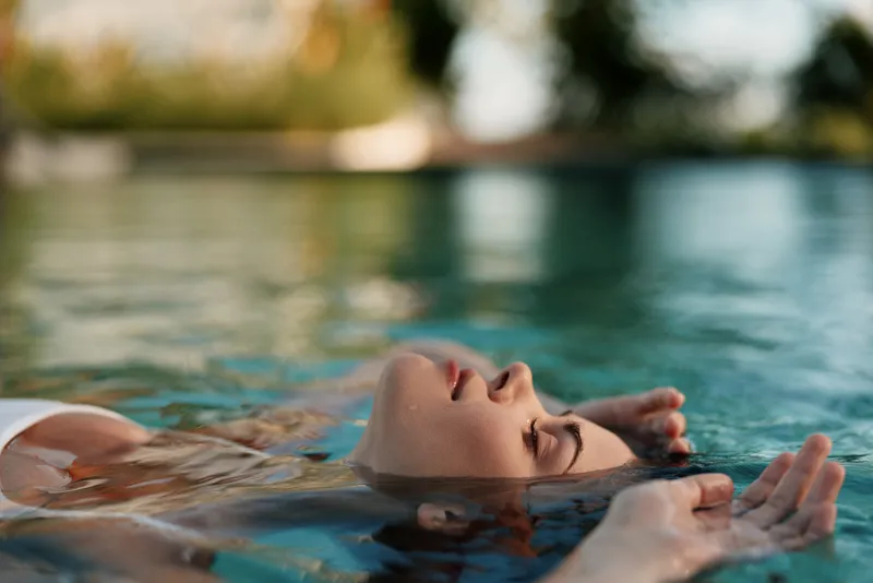 Relaxed woman floating in a pool with eyes closed, sunlit water and soft breeze, tranquil mood, clear blue surroundings, casual summer vibe, carefree lifestyle sun drenched horizon warm tones