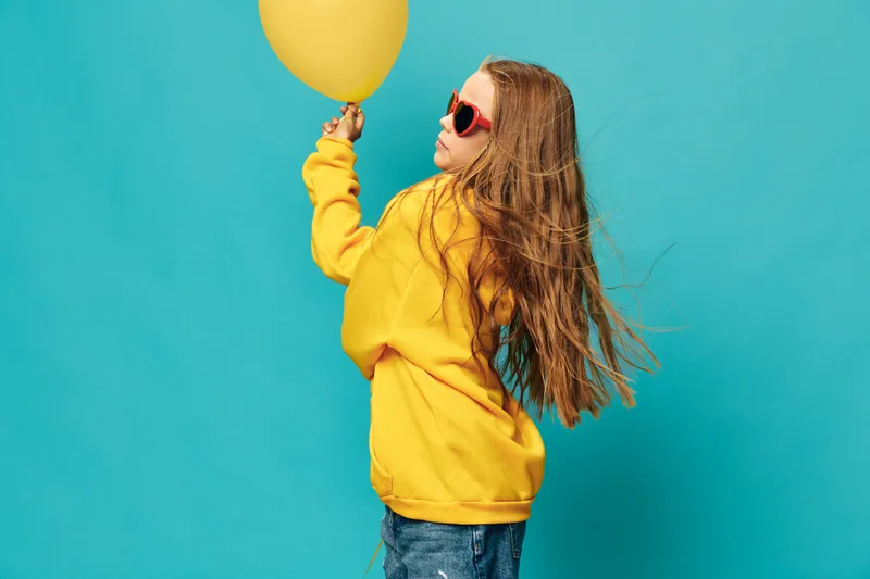 Girl with long hair in yellow hoodie and jeans holding yellow balloon against blue background. Child wearing sunglasses looking upward with playful and joyful mood in studio lighting.