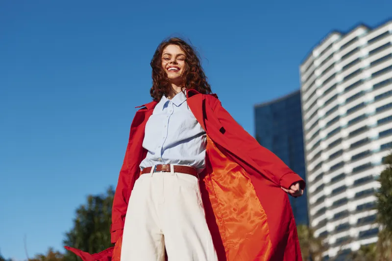 Smiling young woman with curly hair wears a bright red coat over a light blue shirt and white trousers, standing outdoors with modern buildings looming behind her on a sunny day.
