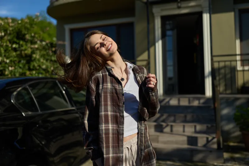 A woman basks in bright sunlight, hair tossed by a breeze, wearing a plaid shirt over light clothing. Concept for casual, carefree female lifestyle moments