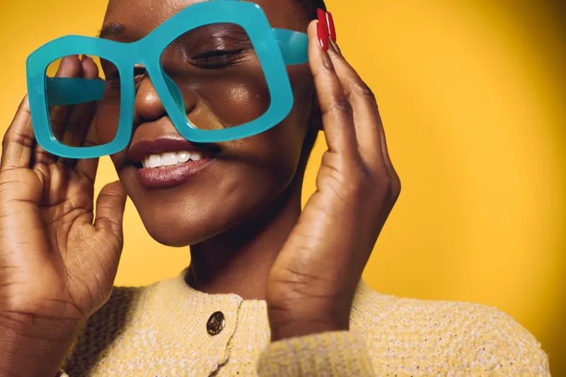 Close-up of a smiling black woman with dark skin wearing large blue glasses and a beige sweater, posing against a bright yellow background. Concept of fashion, joy, and confidence.