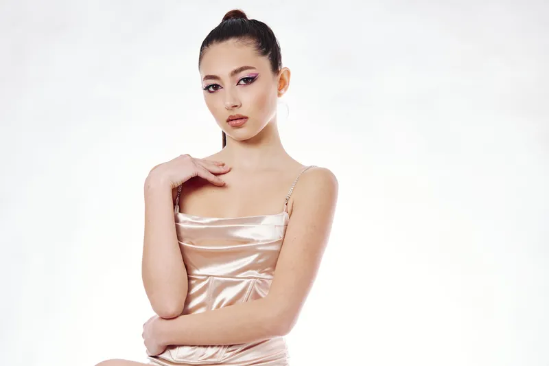 Elegant young woman with slicked back hair in a shiny satin dress poses confidently against a white background. Styled makeup highlights eyes with purples and strong brows.