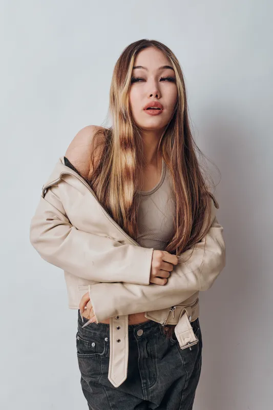 Woman with long hair wearing beige jacket and tank top posing against white wall. Casual fashion outfit with neutral colors and relaxed confident posture in studio setting.