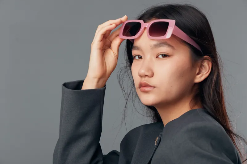 Young woman with long dark hair and serious expression wearing pink sunglasses and gray blazer posing on gray background in studio for fashion photography portrait.