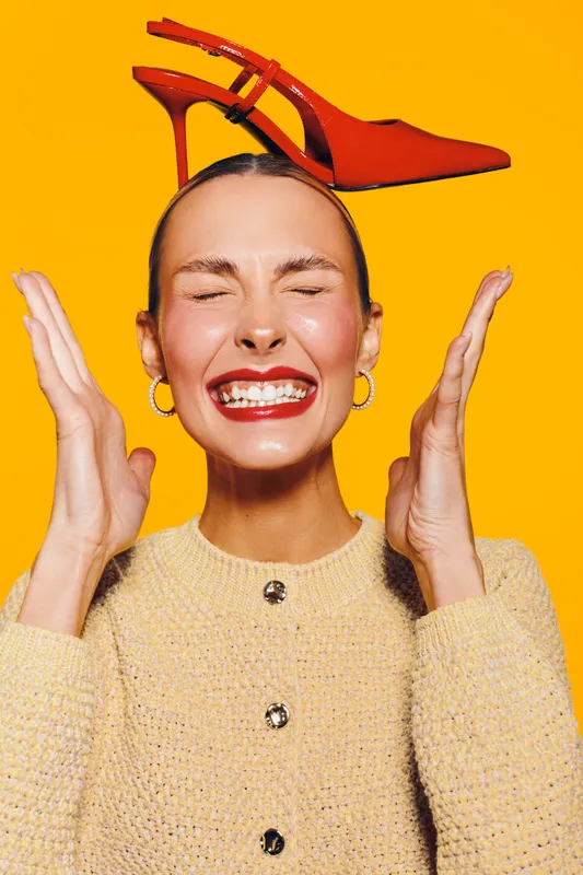 Young woman with red high heel shoes on her head smiles with closed eyes and raised hands. Bright yellow background and beige sweater create a playful fashion concept.