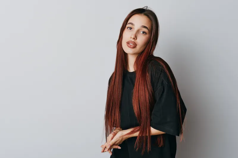 Young woman with long red hair and casual black t shirt posing and looking at camera with neutral expression on gray background in studio. Female model with natural makeup and straight hair.