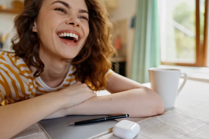 Smiling woman with wavy hair leans on a desk in a cozy home, writing with a pen beside a white mug, radiating warmth and relaxed focus.
