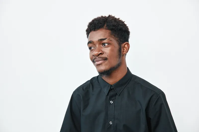 Young black man with dark skin wearing black shirt looking aside with thoughtful expression on white background. Portrait of African ethnicity male with casual style and short hair.