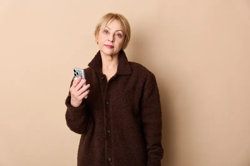 Woman holding smartphone in studio portrait against beige background, mature woman using technology for communication, soft lighting and calm mood, copy space