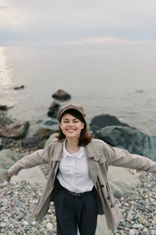 Happy young woman in casual clothes and cap stands with arms outstretched on rocky shore near calm sea under cloudy sky. Concept of freedom, joy, and nature connection outdoors.