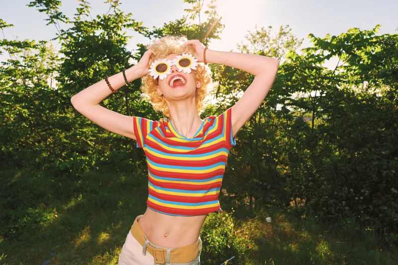 Young woman outdoors laughs with flower sunglasses and striped shirt, enjoying a sunlit moment amid lush greenery and warm summer breeze.