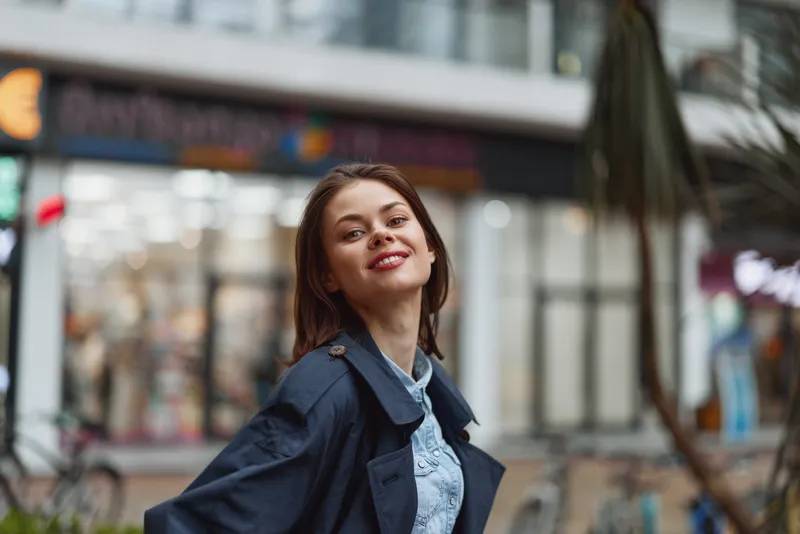 Young woman with a bright smile stands outdoors on a city street, wearing a denim jacket and light shirt, candid urban portrait with blurred storefronts.