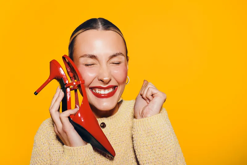Happy woman with closed eyes holds red high heel shoes near face, smiling with excitement on bright yellow background. Concept of fashion, joy, and stylish footwear.