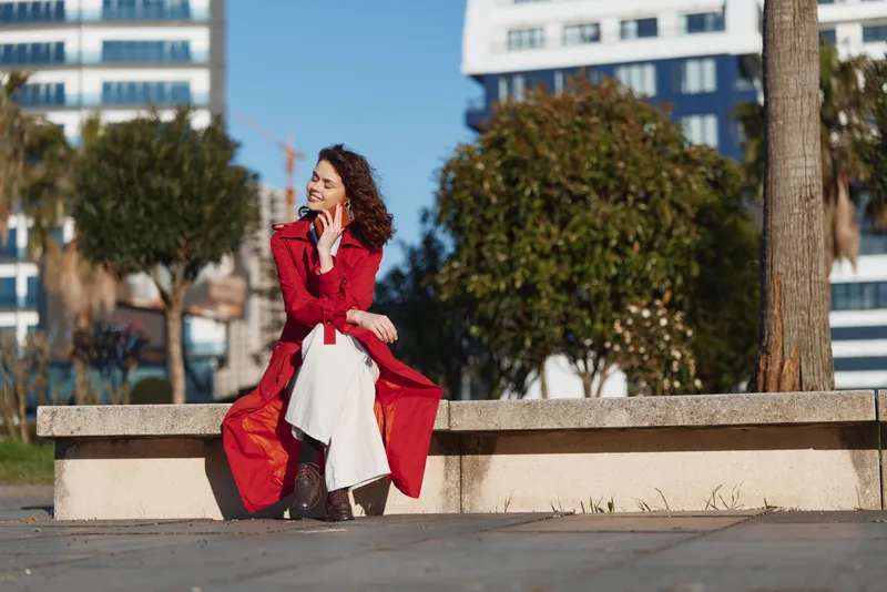A woman in a red coat sits on a concrete bench in an urban park, talking on a phone, with a white dress peeking out, a sunny day, calm and stylish.