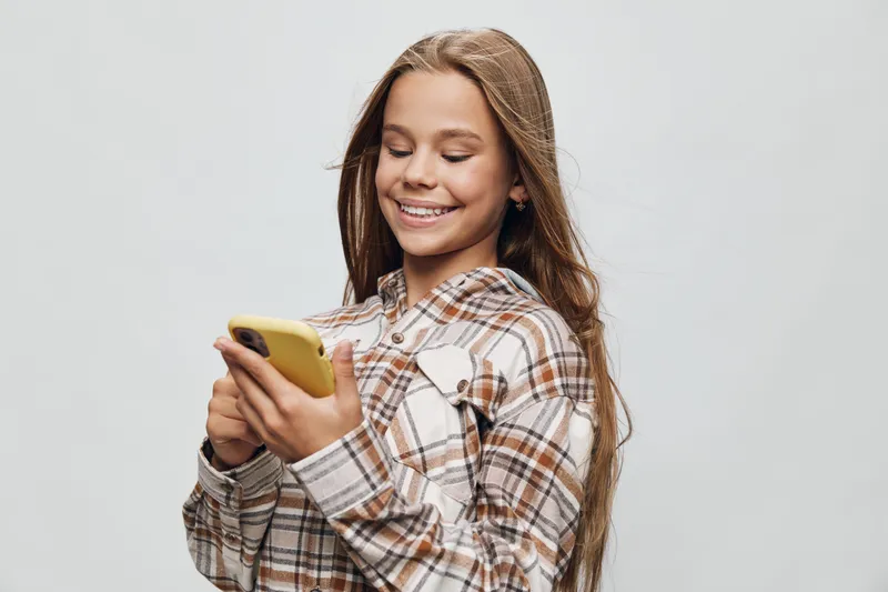 Smiling girl holding yellow phone browsing or texting with casual plaid shirt looking down on screen with long hair on light background for social media or communication.