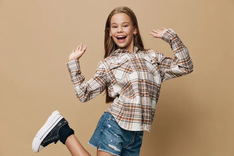 Girl smiling and jumping with raised hands in casual plaid shirt and denim shorts on beige background. Happy child wearing sneakers and enjoying energetic pose of fun and youth.