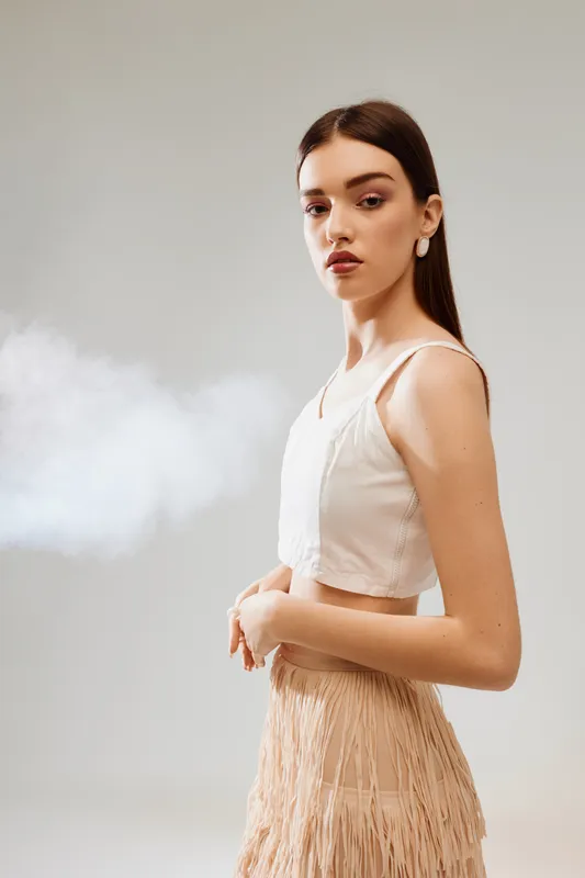 Young woman in beige top and fringed skirt poses confidently in studio with soft lighting. Concept of beauty, fashion, elegance, and modern style.