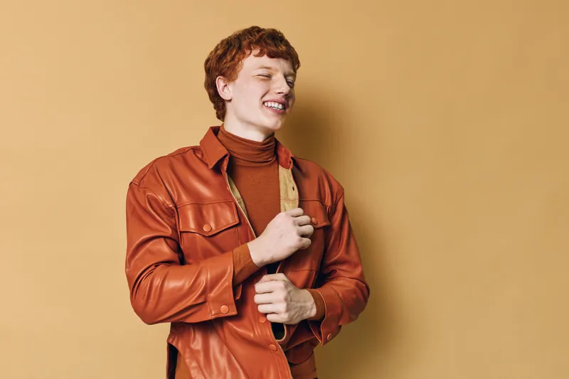 Young man with red hair smiling and eyes closed wearing brown leather jacket and turtleneck sweater against beige background in casual fashion portrait indoors.