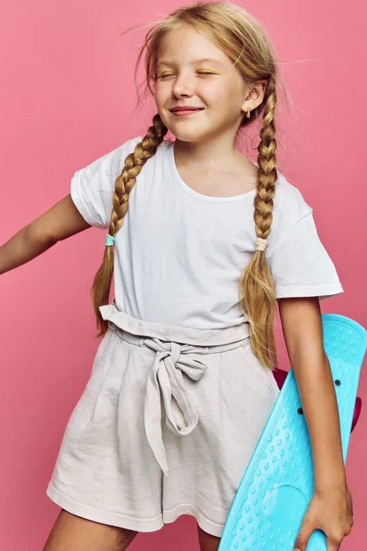 Happy young girl with braided hair stands against pink background holding turquoise skateboard. She wears a white t-shirt and beige shorts with eyes closed, smiling gently. Concept of childhood,