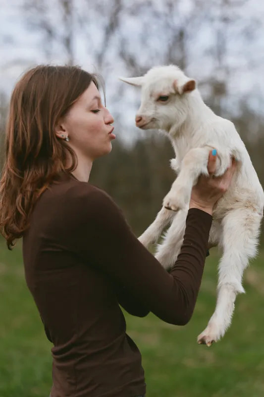 Cheerful woman outdoors lifts a playful goat kid, sharing a gentle kiss in a sunlit field, natural colors, relaxed pose, soft breeze, countryside atmosphere, animal bond
