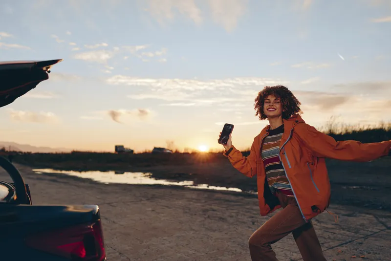 Woman lifestyle outdoors beside car at sunset, running on road in orange jacket, curly hair smiling, holding smartphone, golden hour light, scenic landscape