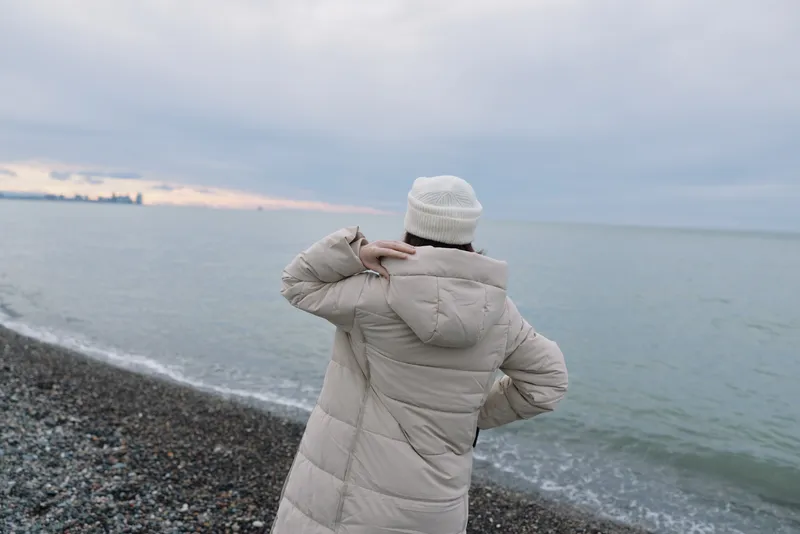 Winter coast sea beach person standing in a warm jacket and beanie, gazing at the calm horizon along the coastline, a quiet moment by the sea.