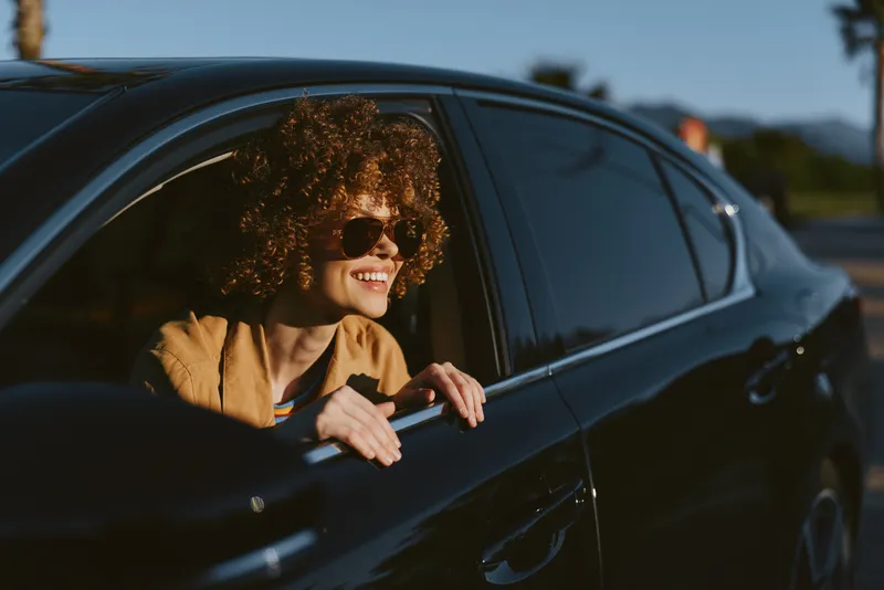 Young woman with curly hair leans out of a sunlit car window, smiling as a warm breeze lifts her hair and brightens the moment