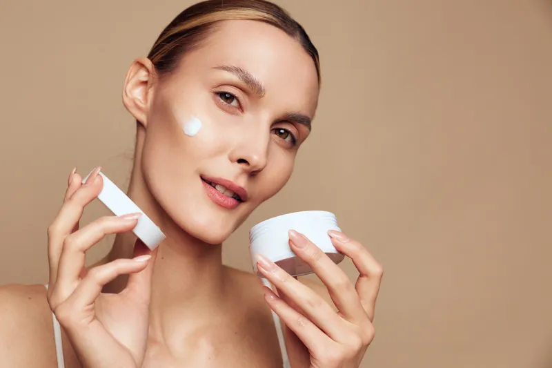 Young woman applying moisturizing cream on face, healthy skin care routine, natural beauty, soft lighting, neutral background, close up portrait, skincare concept