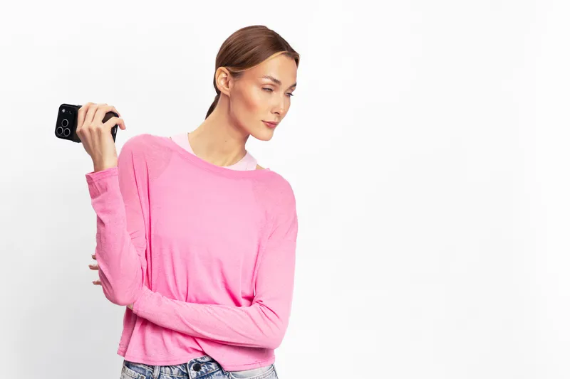 Young woman in pink sweater holding a smartphone looks away with a thoughtful expression. Isolated on white background. Concept of communication, technology, and casual fashion.