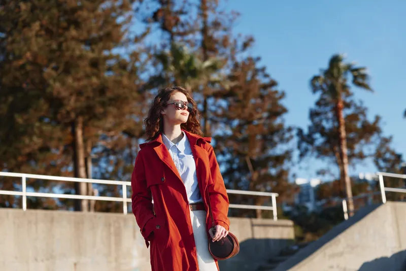 Young woman in a red coat walks outdoors with sunglasses, posing confidently on a sunny day amid trees and concrete structures, her gaze distant and hands in pockets, showcasing urban chic and self