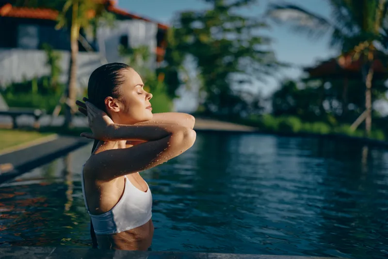 Poolside scene with a woman resting at the edge, eyes closed in quiet relaxation. Warm sunshine on clear water in a tropical resort suggests escape, wellness, and calm.