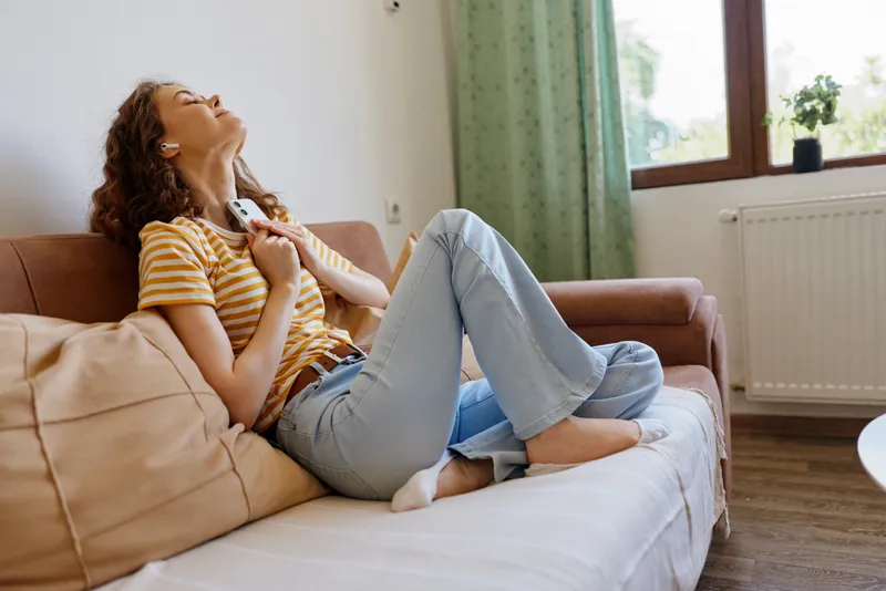 Cheerful woman at home, relaxing on a sofa, striped top and jeans, sunlit living room, gentle breeze through window, natural colors, cozy and carefree moment