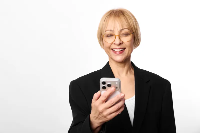 Woman smiling holding smartphone, glasses, blonde hair, business portrait on white background, communication and technology, friendly casual mood today, senior energy