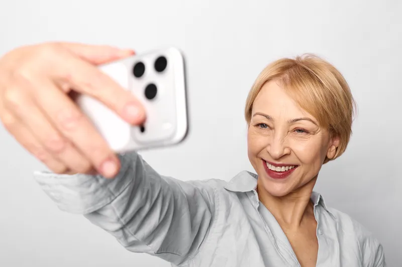 Woman selfie smartphone, laughing senior indoor, holding a phone at arm length, close up portrait on a gray background, candid joy, soft studio lighting, lifestyle technology moment
