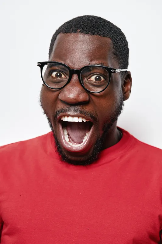 African American man with dark skin and glasses wearing red shirt expressing surprise or excitement with wide open mouth on white background. Closeup portrait of emotional black male.