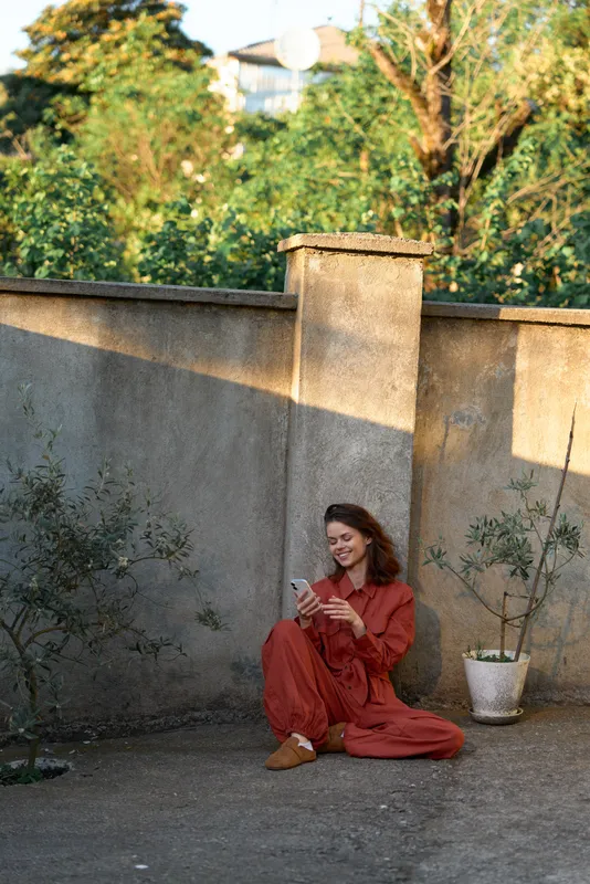 Young woman in a red dress sits on the ground beside a textured wall in a sunlit courtyard, scrolling on a smartphone with a calm, focused expression under warm shadows and distant greenery