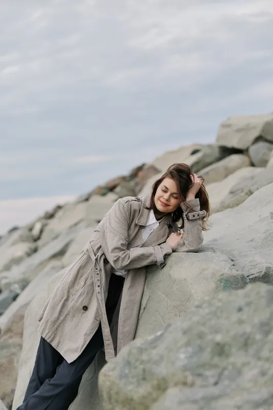 Young woman in knitted sweater leans on large rocks outdoors with closed eyes. Relaxed mood in nature with cloudy sky background. Concept of calm, comfort, and autumn fashion.