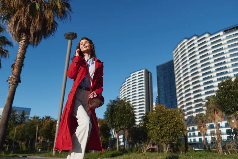 Young woman in a red trench walks through a sunny cityscape with modern high rises, smiling while talking on her phone and enjoying a breezy day outdoors